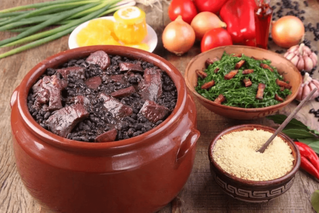 圖片展示了一道巴西傳統料理——黑豆燉肉（Feijoada）。一個棕色陶罐裝著濃郁的黑豆燉肉，旁邊還有配菜，包括綠色的蔬菜、玉米粉，以及切片的橙子和其他蔬菜。這道菜通常以豐富的黑豆、豬肉和香腸為主料，並搭配米飯食用，是巴西的代表性美食之一。